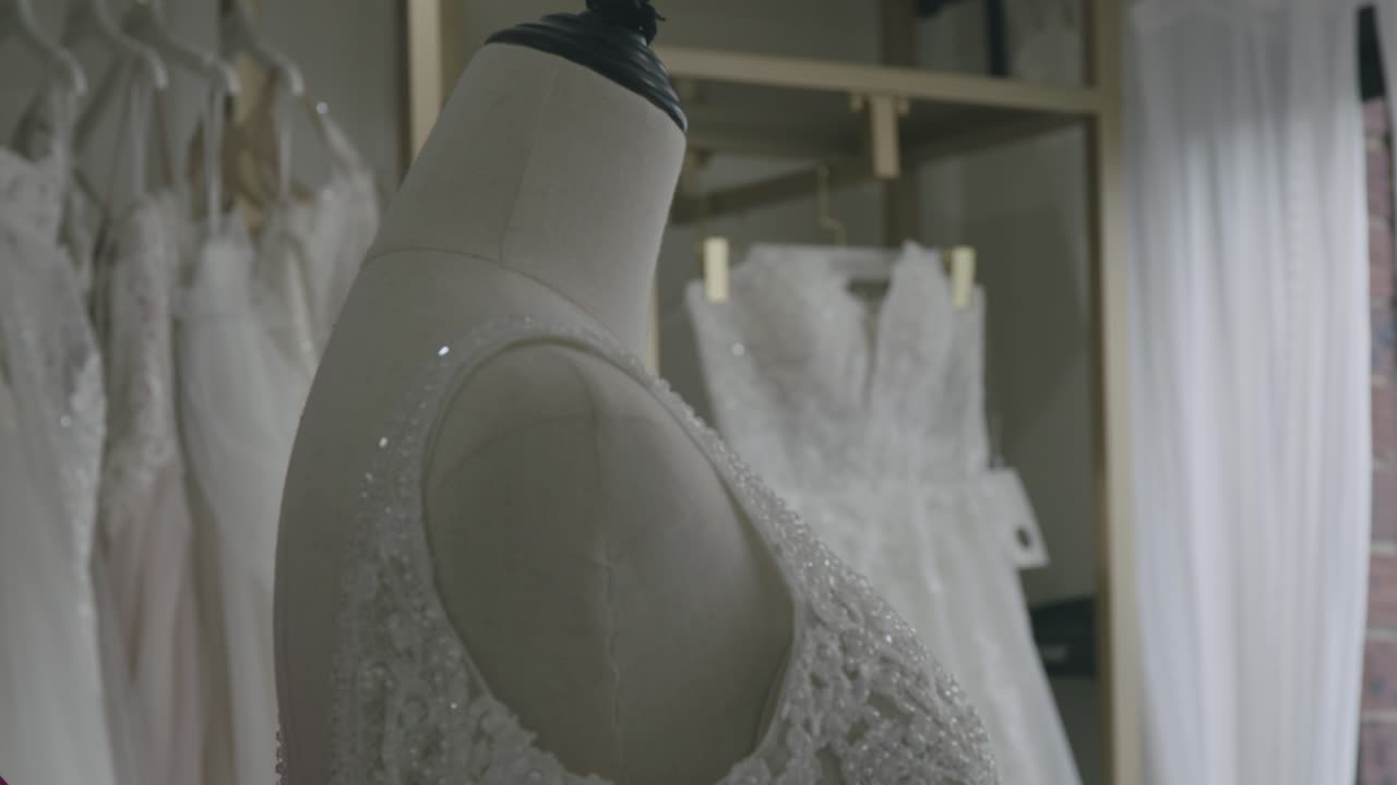 An Asian woman in bright lighting adjusts a delicate, beaded wedding gown in a bridal store. The focus centers on intricate fabric details, creating an intimate, elegant atmosphere