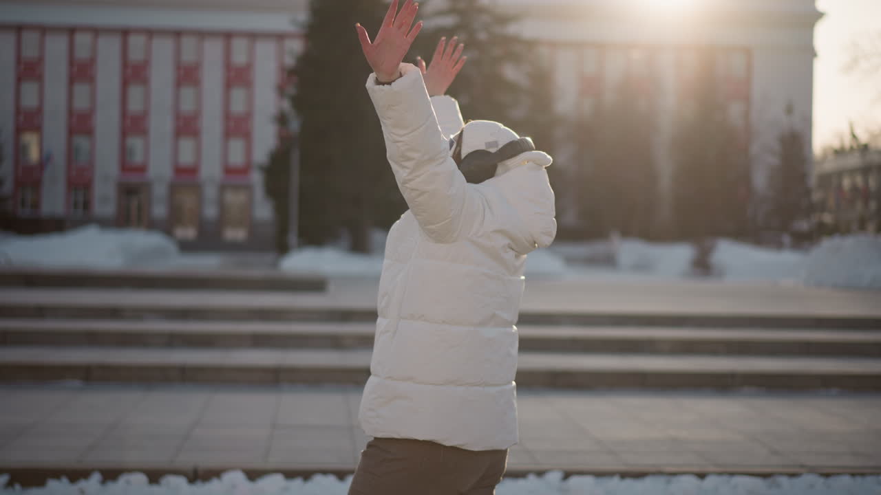 Side view of creative woman expressing herself through joyful motion on city steps during cold winter evening as sunlight gently backlights her silhouette in rhythmic dance wearing white coat