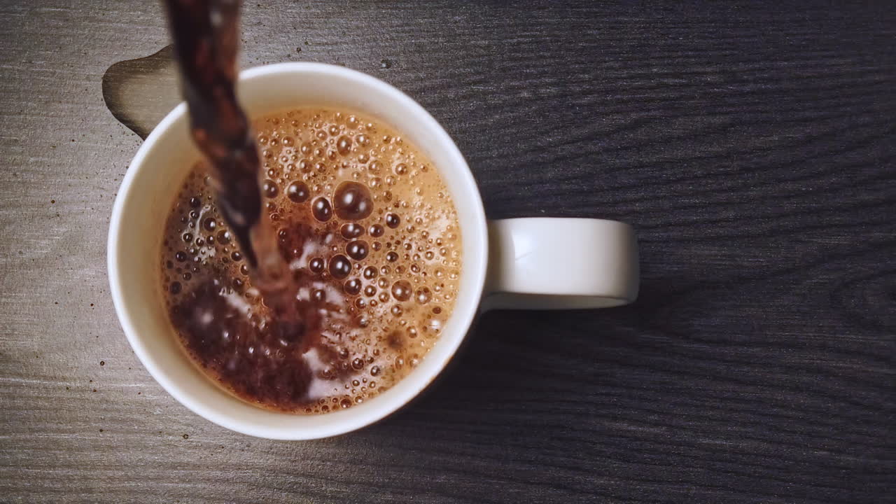 Slow motion-optagelse af cola, der hældes i en hvid keramikkop på et træbord i køkkenet