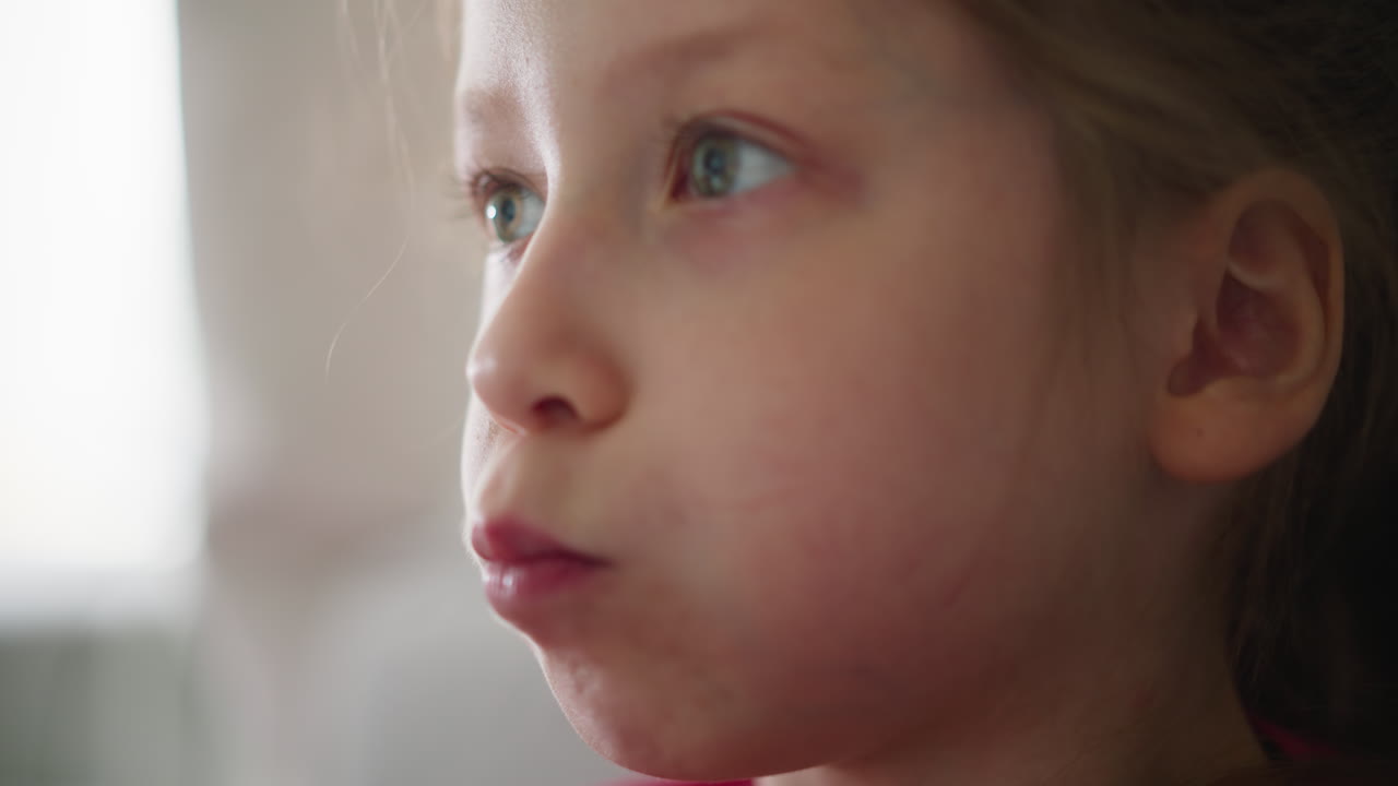 Close up of kid in red top savoring snack as she chews with focused expression and natural lighting, enjoying delicious moment against soft bright blur background