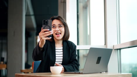 Pretty smiling lady with short dark hair and glasses looks at her smartphone. Woman having online video chat on her gadget.