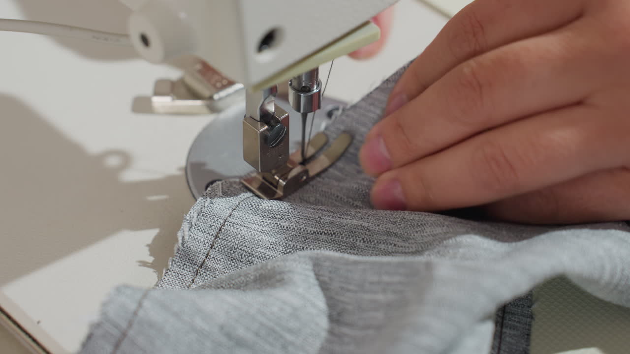 Close up of sewing machine needle stitching light gray fabric while hand steadily guides material through presser foot during garment production in bright workspace, showing precision of textile work