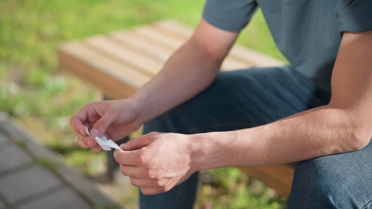 partial view of young man seated on wooden bench outdoors holding and unwrapping nicotine patch under sunlight with green grass and pavement in background casual clothing and focused gesture visible