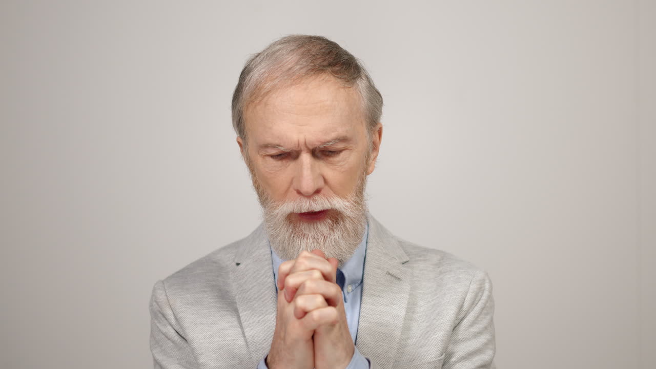 Uomo stressato che prega al chiuso. Anziano triste che si stringe le mani su sfondo da studio.