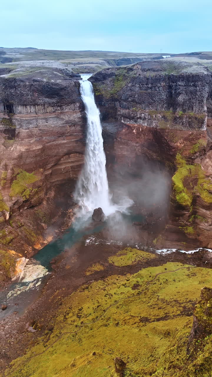 Approaching a couple standing on the edge of the dangerous rock. Drone flies to the chute in the mountains revealing the view on the splendid waterfall and river flowing among the rocks. Iceland. Vertical video.