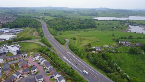 aerial video of scottish moterway countryside, freeway with traffic surrounded by green vegetation and buildings on one side and lakes of water