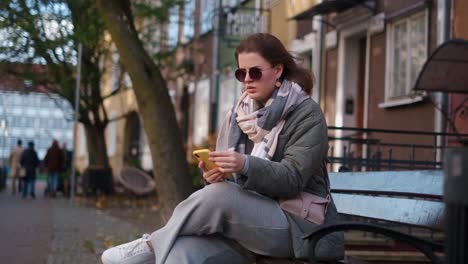woman sitting on a bench, using a smartphone in an autumn city street