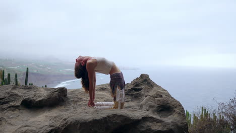 Amidst-the-azure-sea-and-blue-sky,-a-girl-does-yoga-on-the-rocks,-assuming-a-bridge-posture-while-standing-on-a-stone