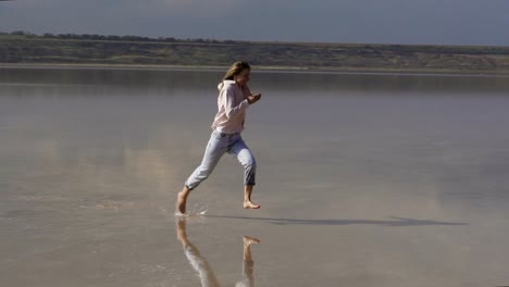 Eine-Glückliche,-Langhaarige-Frau-In-Freizeitkleidung-Läuft-Im-Sommer-Auf-Dem-Seichten-Sandstrand-Des-Sees-Und-Lächelt