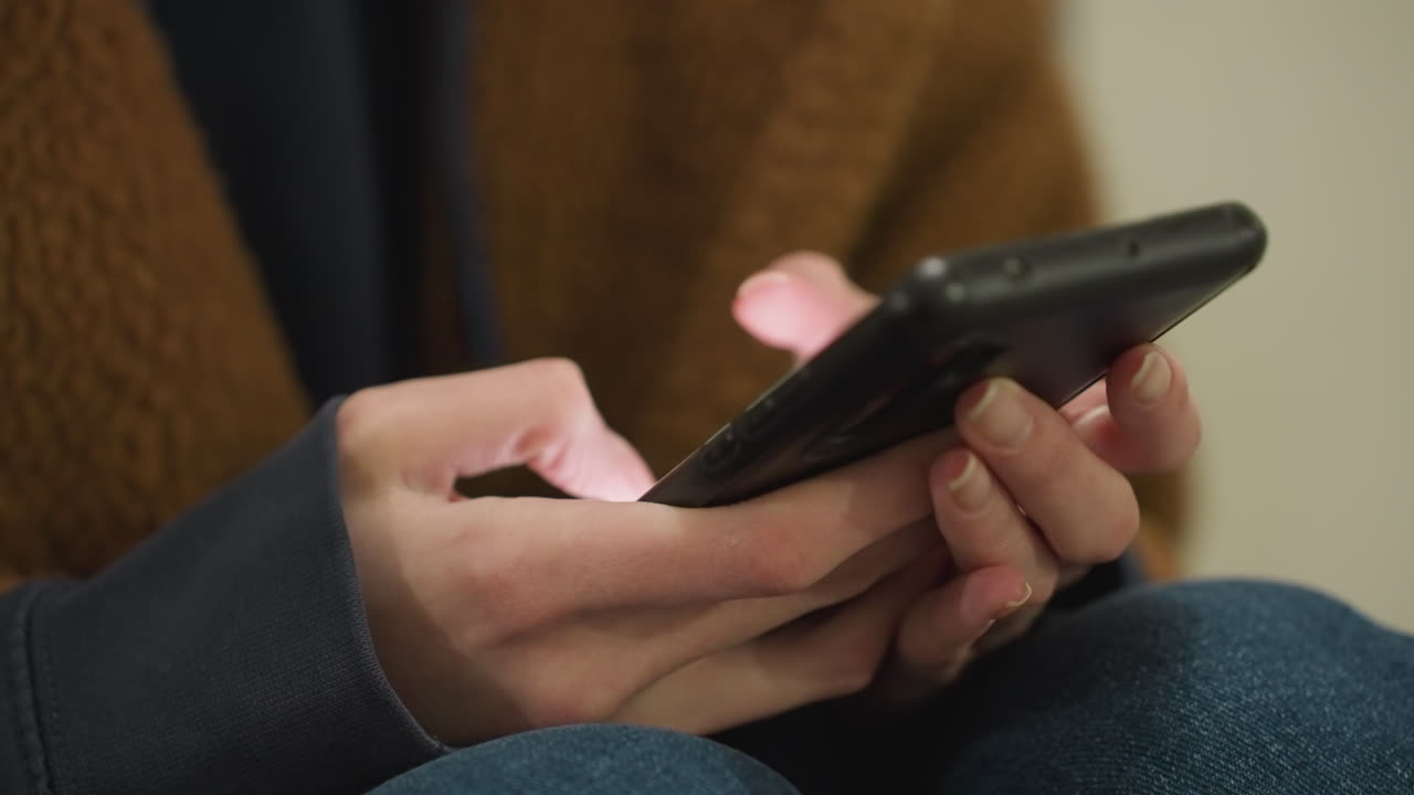 primer plano de manos con abrigo marrón y vaqueros azules, sosteniendo y presionando un teléfono inteligente con ambas manos. la interacción con la tecnología, enfatizando el enfoque en el dispositivo en un entorno urbano