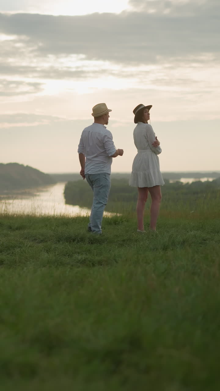 모자,  셔츠, 청바지를 입은 남자가  드레스를 입은 여자를 향해 걸어 가며, 해가 지는 동안 호수를 내려다보는 잔디 언덕에 서 있습니다.