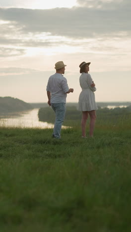 un hombre con sombrero, camisa blanca y vaqueros camina hacia una mujer con un vestido blanco de pie en una colina cubierta de hierba con vistas a un lago al atardecer. momento con un hermoso paisaje