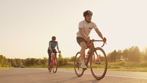 Un-Hombre-Y-Una-Mujer-Montan-Bicicletas-Deportivas-En-La-Carretera-Al-Atardecer-Con-Equipo-Y-Cascos-Protectores-En-Cámara-Lenta-A-120-Fps.