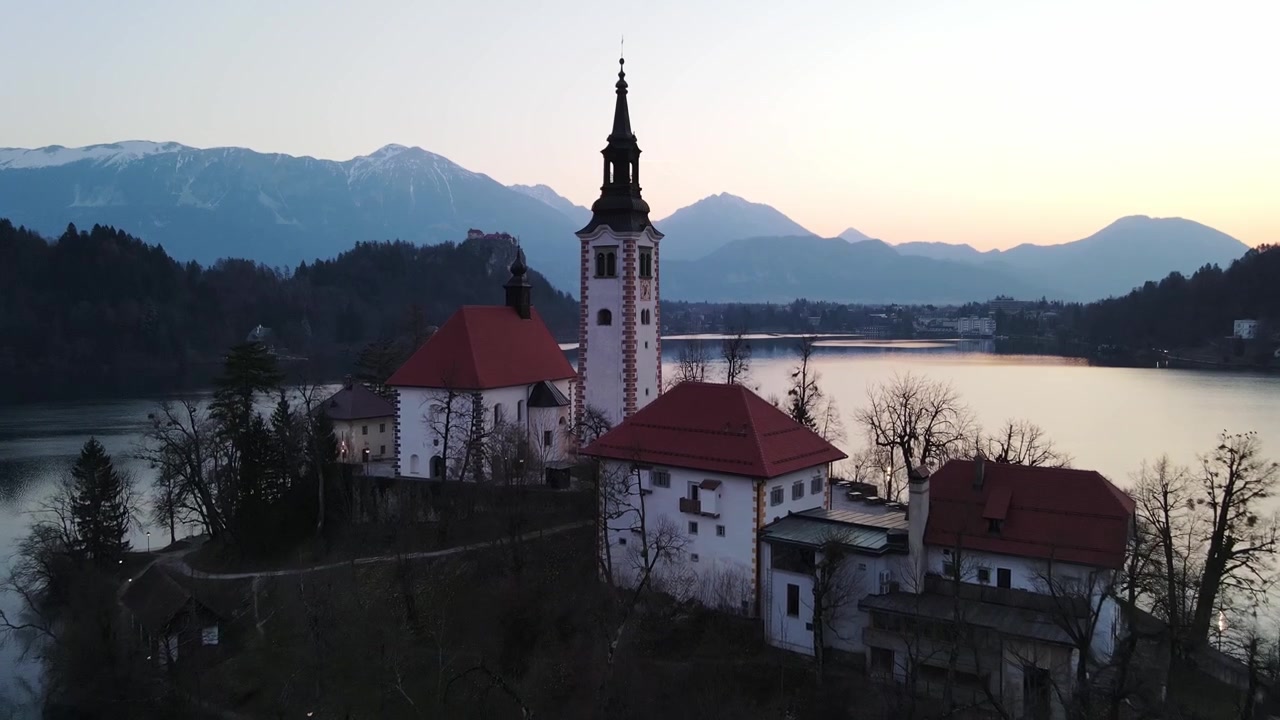Volo di drone verso la Chiesa dell'Isola di Bled all'alba sopra un calmo lago alpino.