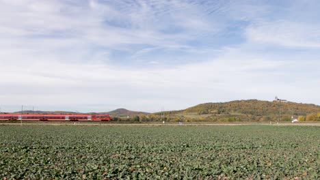 El-Tren-Rojo-Deutsche-Bahn-Pasa-Por-Un-Campo-De-Coles-De-Otoño-Con-Un-Castillo-En-La-Cima-De-Una-Colina-Al-Fondo,-Durante-El-Día