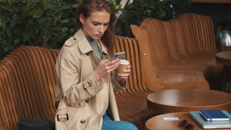 caucasian female student using smartphone and drinking coffee outdoors.