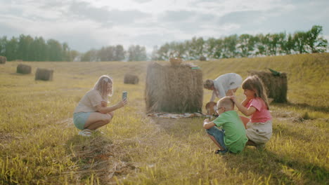 woman crouches to take photo of kids playing while friend spreads blanket as playful dog climbs on it, sunlit countryside setting captures joyful family bonding and outdoor fun