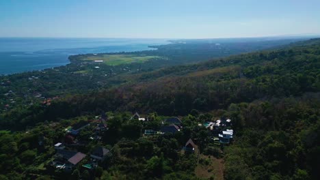 Drone aerial shot over North Bali, capturing the blue sea, village houses, and vibrant green trees under bright daylight, showcasing a serene and warm tropical atmosphere