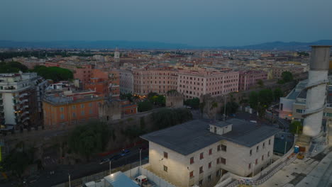Fly-over-ancient-city-walls-dividing-railroad-tracks-and-blocks-of-apartment-buildings.-Low-flight-above-rooftop-terraces-at-dusk.-Rome,-Italy