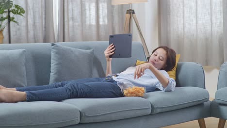 teenage girl relaxing on couch, watching tablet, eating snacks