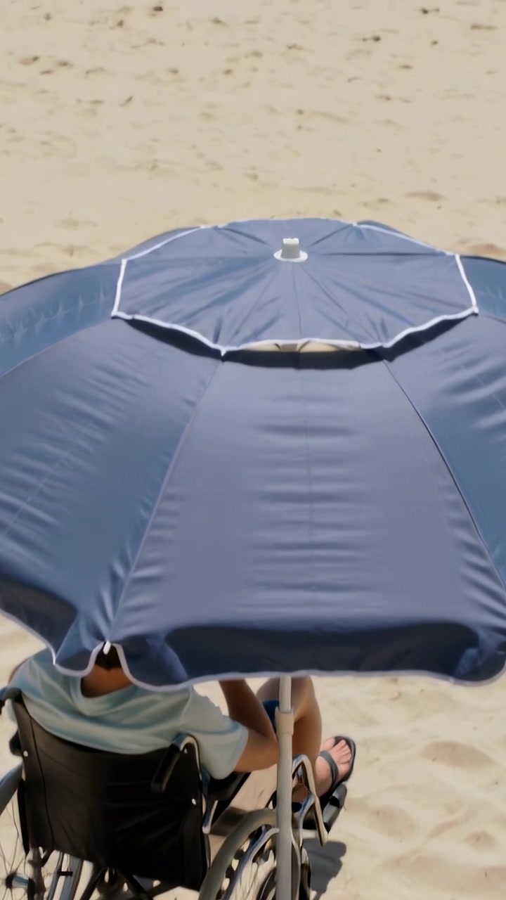 Video verticale: Telecamera che si inclina mostrando un utente in sedia a rotelle in pantaloncini che guarda le onde sotto un ombrellone blu.