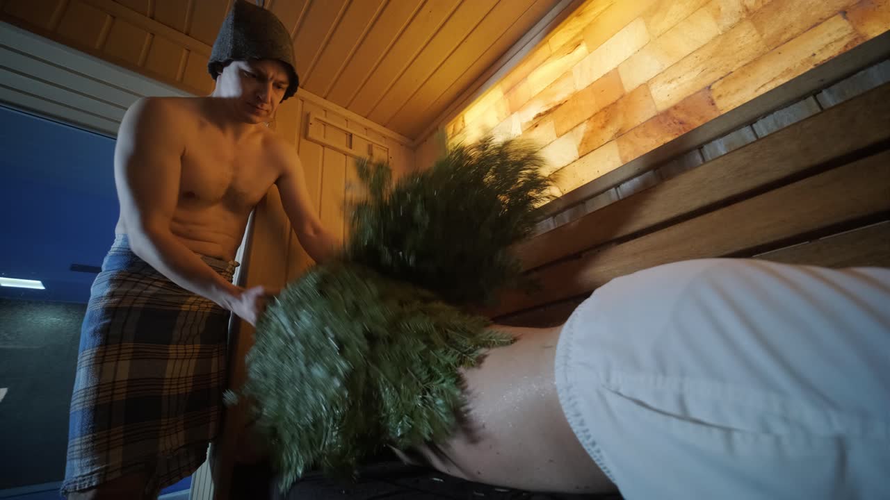 hombre tendido en un banco de madera en el spa con masajista haciendo masaje con escoba de roble. cliente en sauna finlandesa en el interior con profesional.