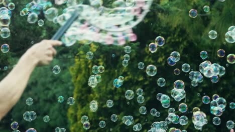 Seifenblasen-Fliegen-Im-Sommer-Draußen-Auf-Einem-Spielplatz-In-Der-Luft,-Eine-Person-Winkt-Mit-Einem-Großen-Runden-Seifenblasenmacher