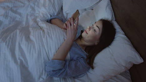 vista de cerca de la encantadora mujer caucásica con cabello oscuro acostado en la cama, charlando en el teléfono inteligente a altas horas de la noche