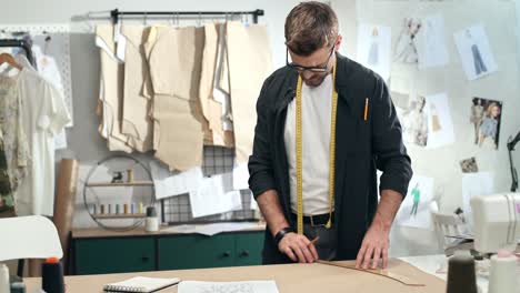 man tailor in eyeglasses taking measurements on table in the sewing workshop and smiling at camera