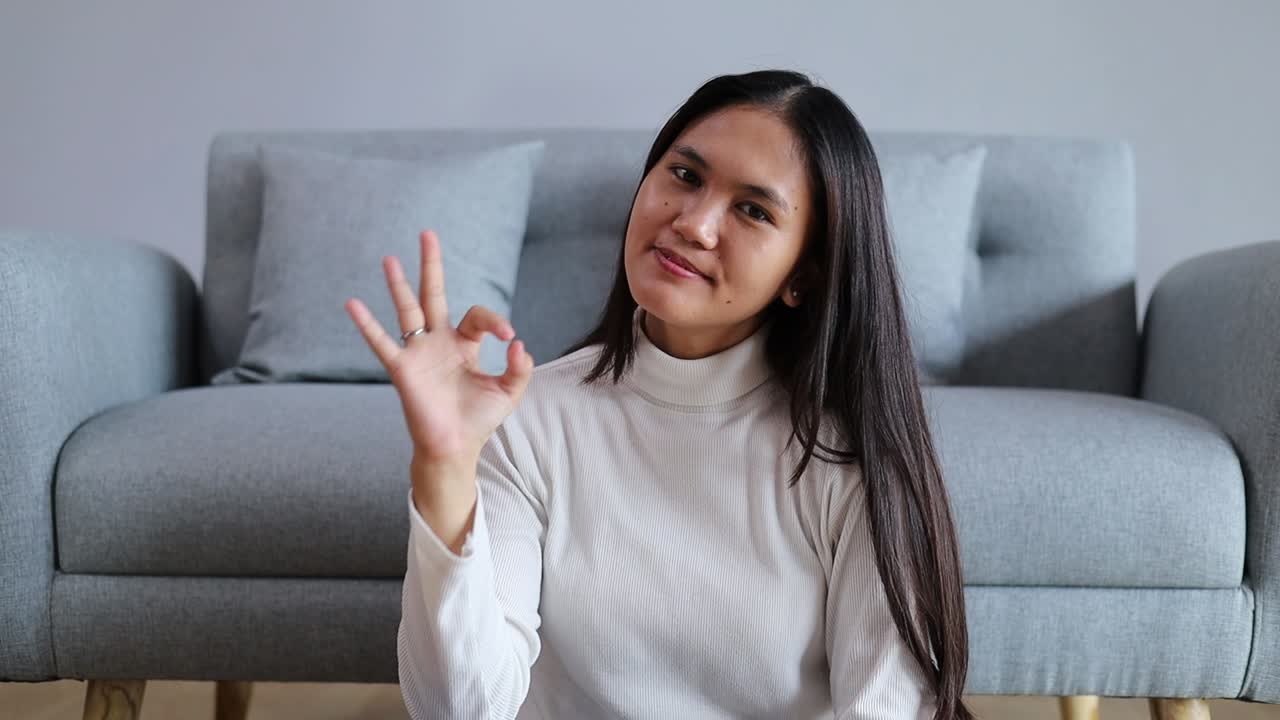 Giovane donna asiatica che mostra il segno OK con la mano, con uno sguardo fiducioso che esprime accordo e un atteggiamento positivo.