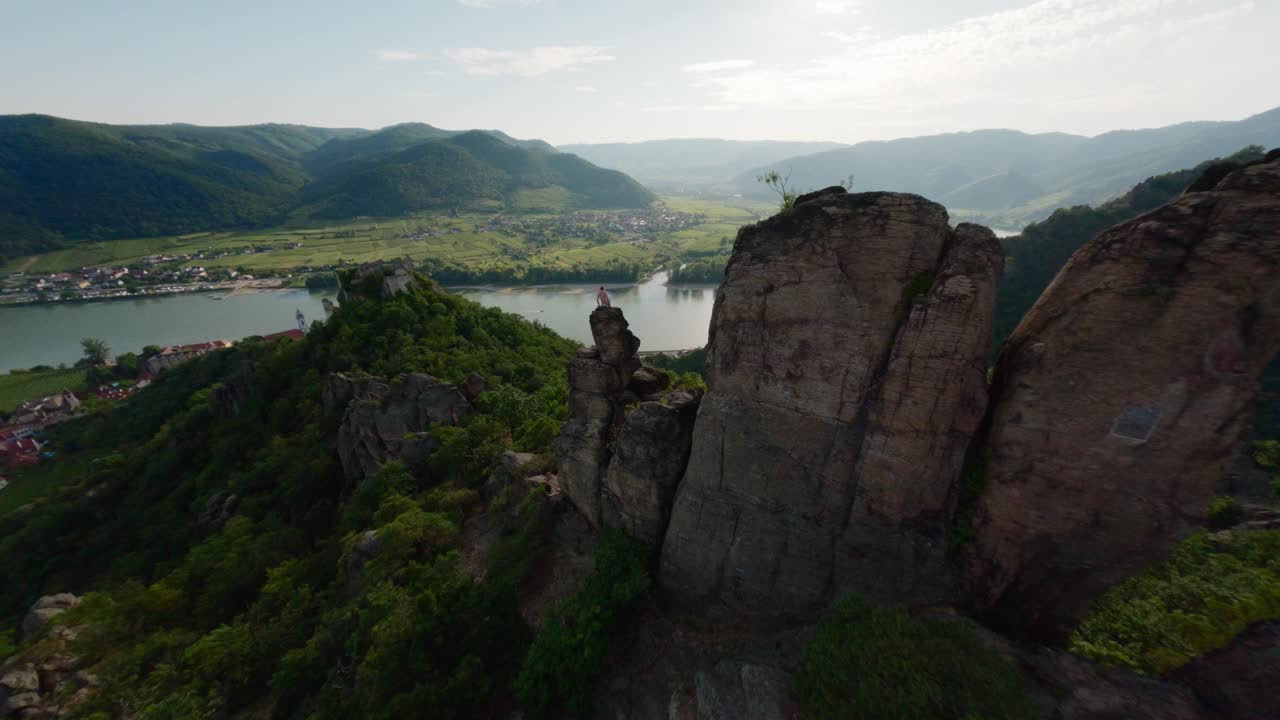 hombre de pie en la cima de una roca de montaña, fpv volador, green valley overview flyover, persona en la cima del mundo, río danubio en el fondo, toma aérea de alta velocidad, vista espectacular cinematográfica, austria