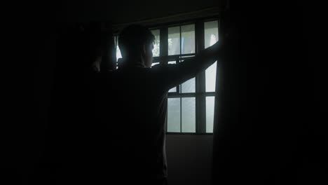 rear static shot of young man opening curtains to let sunlight into dark bedroom.