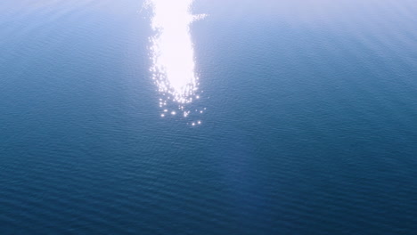 drone flying over a coastline towards a blue ocean with strong sun reflections