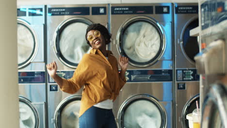 beautiful african american cheerful woman in stylish outfit having fun and dancing in laundry service room while machines washing on background. girl resting while machines working in washhouse.