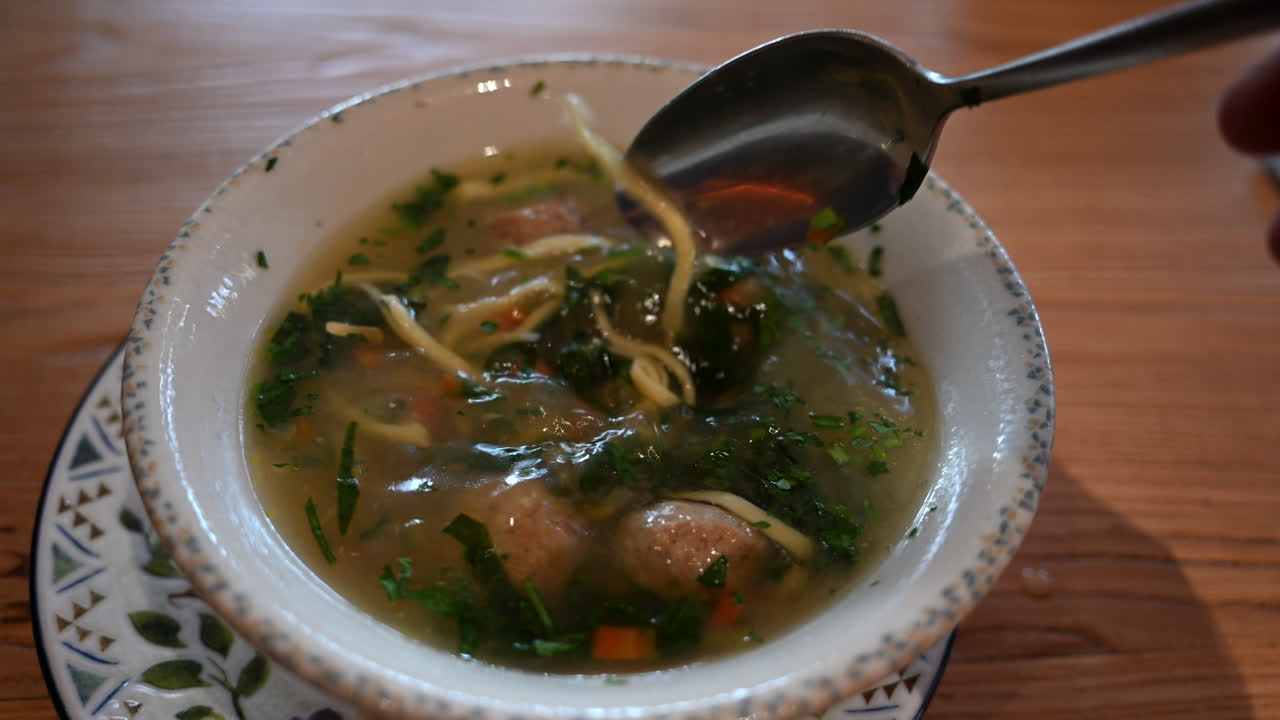 Primo piano di un cucchiaio che solleva noodles e verdure da una zuppa calda e chiara con erbe aromatiche.