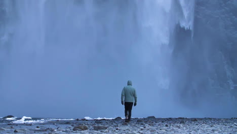 young traveler in blue jacket walking towards huge frozen waterfall in slow motion