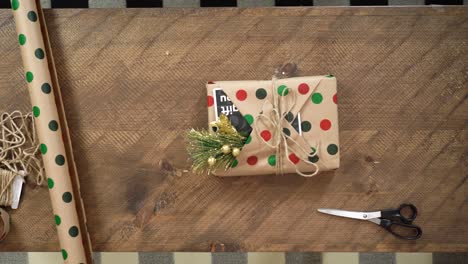 una mujer joven envuelve un regalo de navidad en una mesa de madera con papel de regalo de lunares, una tarjeta y un muérdago listo para poner debajo del árbol de navidad.