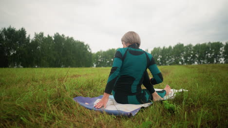 back view of old woman seated on yoga mat in half seated twist, practicing yoga with head slightly tilted backward in a vast grassy field, with trees lined up in the distance under a cloudy sky