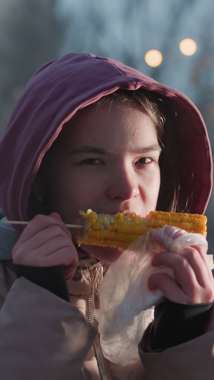 mujer joven disfrutando de maíz fresco en un palo envuelto en una bolsa blanca con vapor que se eleva en un entorno al aire libre, luces bokeh en un fondo borroso, bocadillo de clima frío en la ciudad