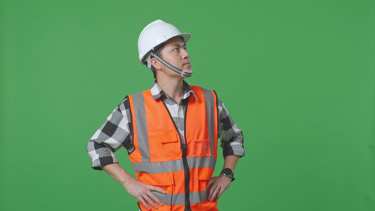 Asian Male Engineer Wearing Safety Helmet Looking Around While Standing With Arms Akimbo In The Green Screen Background Studio