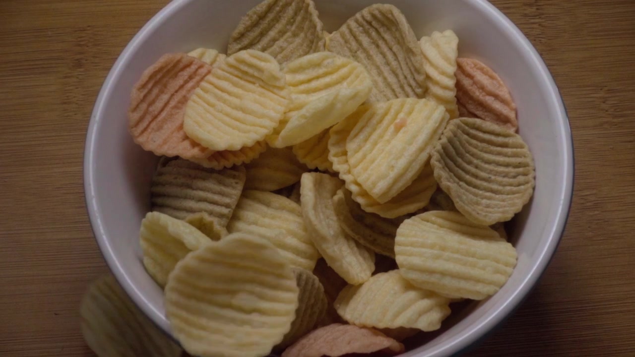 Ridged potato chips falling into white bowl filled with crispy snacks on wooden table showing golden texture and motion during snack preparation