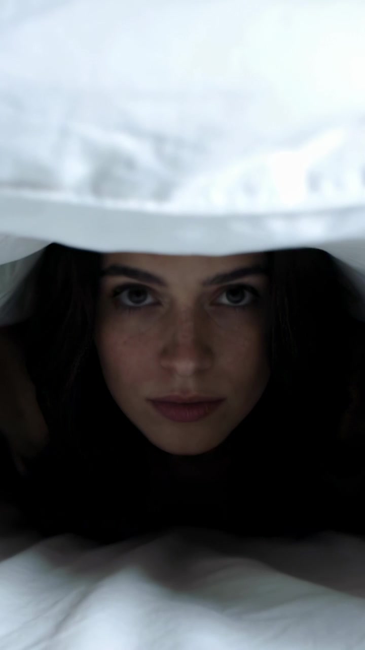 Vertical video: Peeking woman pulling white duvet over face in bed, hiding while wearing dark top