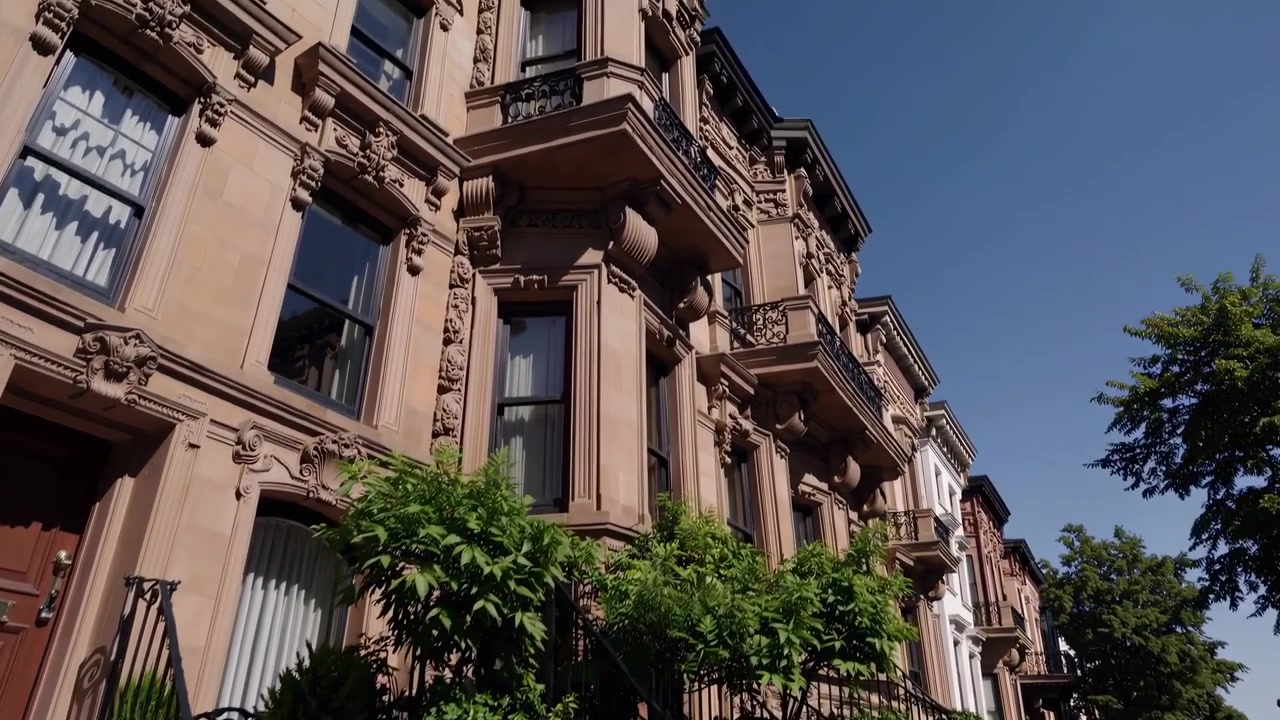 Udsmykkede brownstone-bygninger på en solrig bygade med blå himmel.