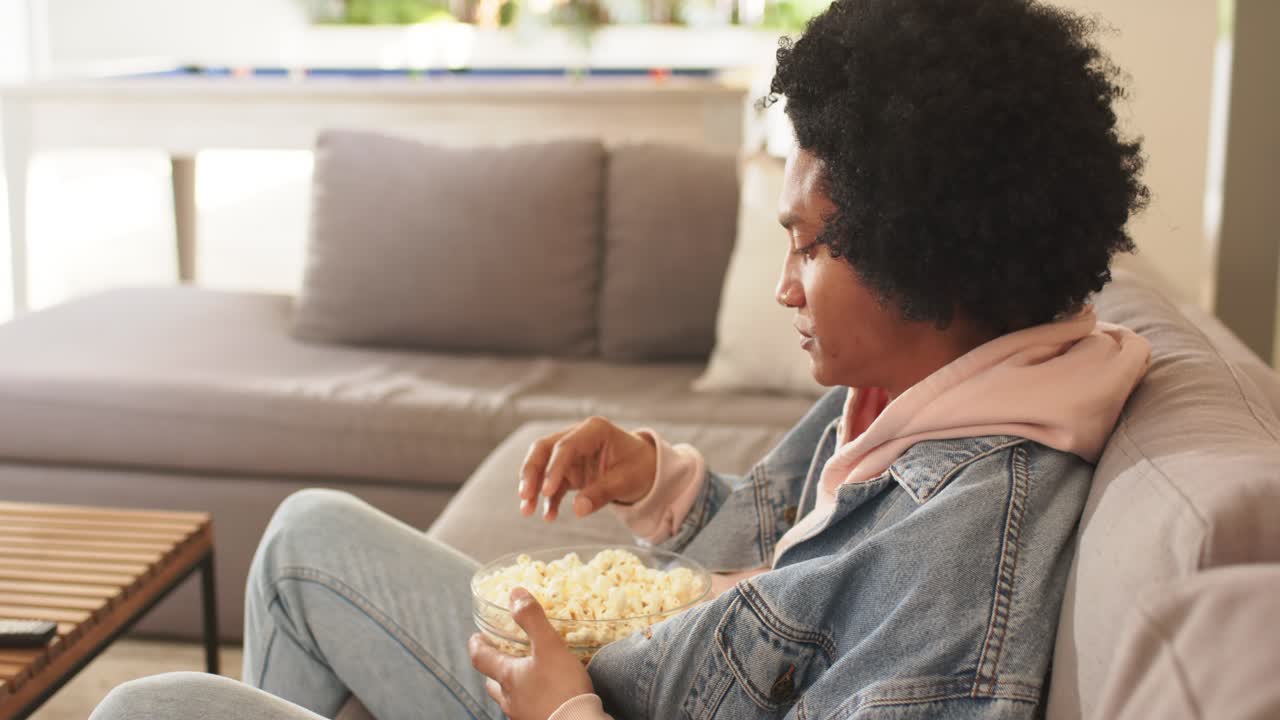 Uomo afroamericano di mezza età guarda la televisione e mangia popcorn da una ciotola sul divano, spazio per il testo