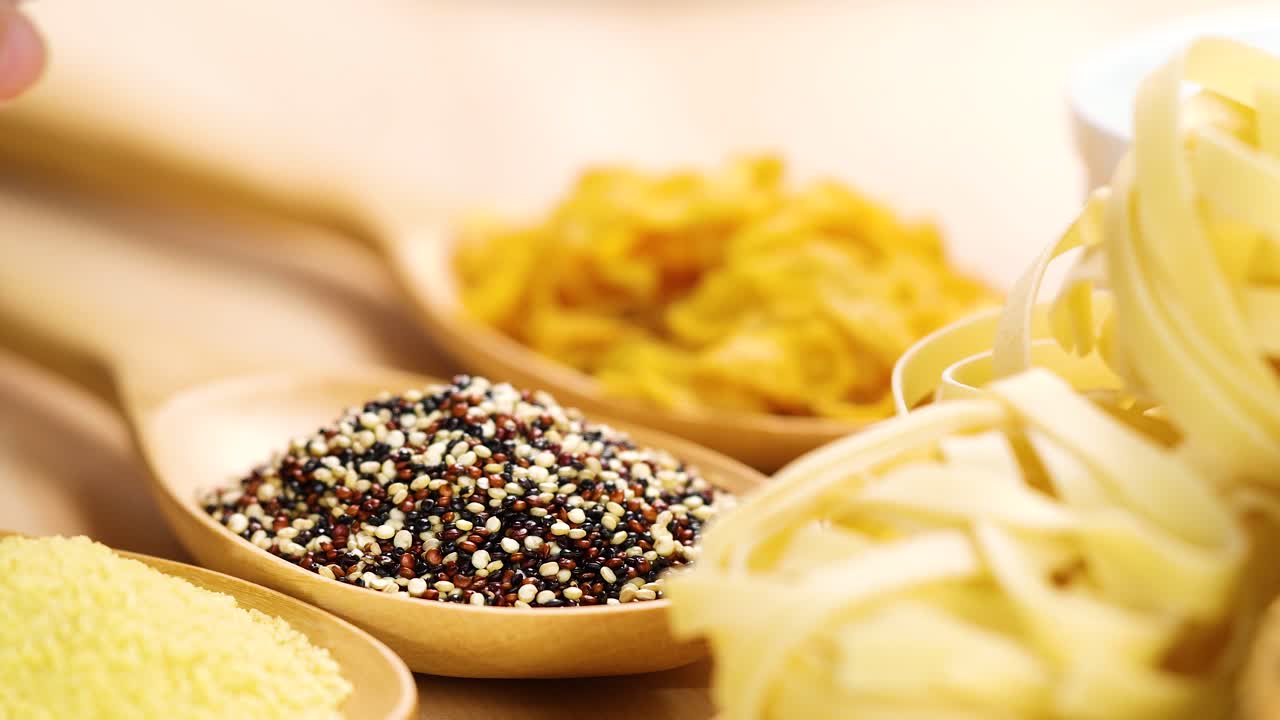Quinoa grains are poured onto a wooden spoon surrounded by pasta and grains, in a warm, well-lit kitchen environment