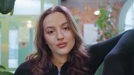 confident woman with long wavy hair leans on cushion and gazes directly into camera in cozy indoor space filled with plants and natural light, evoking calm and self-assurance in modern lifestyle scene