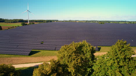 aerial view flying across rural farmland above treetops overlooking solar panel array and spinning wind turbine