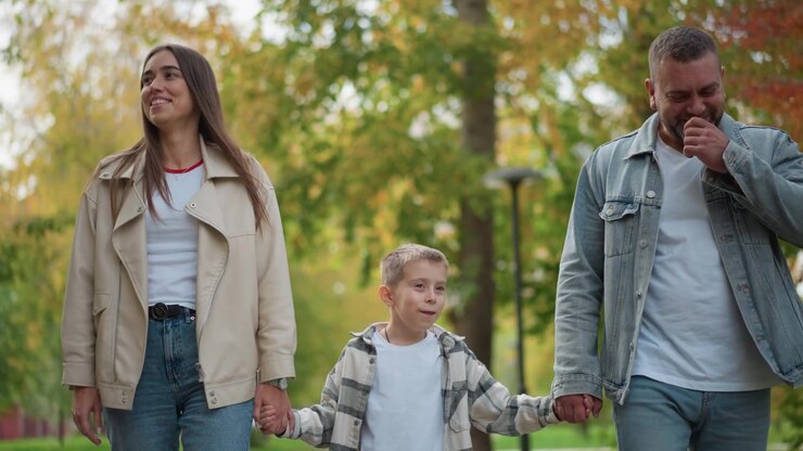 Primo piano di una famiglia che cammina insieme in un parco all'aperto, con la donna e l'uomo che si tengono per mano ai lati del loro bambino. La scena presenta abbigliamento casual, incluse giacche e jeans.