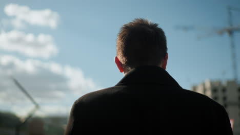 back view of man in black coat engaged in phone call outdoors, shaking head in displeasure while reacting to intense conversation, with urban background including modern buildings, greenery