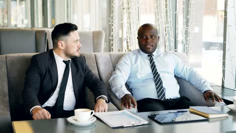 two multi-ethnic businessmen discussing startup ideas in glassy cafe during lunch time. coffee cup, financial graphs and digital tablet are on table